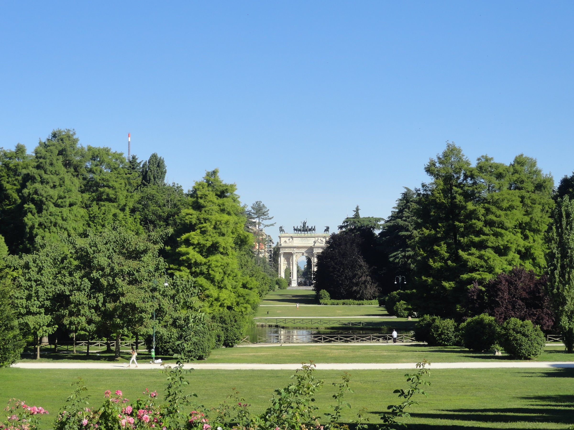 Tree-lined grass avenue in a park leading towards a distant triumphal arch under a clear blue sky.